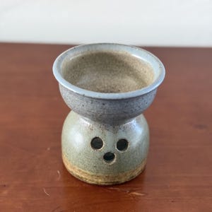 May include: A small, speckled ceramic oil burner with a wide, flared top opening and a rounded base. The exterior features a muted sage green glaze with a light blue rim, and the lower section includes three small, circular ventilation holes.