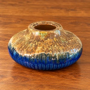 May include: A low, wide ceramic vase with a rounded shape and a small circular opening. The upper portion features a textured, mottled brown and tan glaze, while the lower section has a deep blue glaze with vertical ribbed detailing.