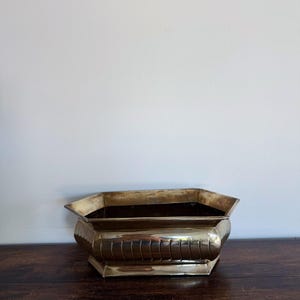 May include: A polished brass planter with a hexagonal shape and a wide, flared rim. The exterior features a series of vertical, curved indentations along the sides, creating a textured pattern. The base is flat and follows the hexagonal form.