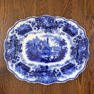 May include: An oval ceramic platter with a scalloped edge, featuring a blue and white transferware pattern. The central scene depicts a castle, mountains, and figures near a bridge, surrounded by a decorative border of floral and scroll motifs.