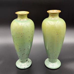 May include: Two ceramic vases with a tapered silhouette and a pedestal base. The vases feature a glossy glaze in shades of light green and sage, decorated with a speckled pattern that transitions into a denser, darker green texture at the base.