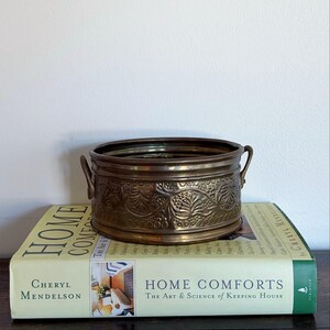 Op de afbeelding: Een ronde, metalen koperen kom met een reliëf bloemenpatroon aan de buitenkant en twee kleine handvatten aan de zijkant. De kom staat op een dik, lichtgekleurd hardcover boek met de titel Home Comforts: The Art & Science of Keeping House.