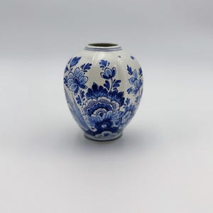May include: A small, bulbous ceramic vase with a white base and intricate blue floral patterns painted across the surface. The vase features a narrow, circular opening at the top and a rounded body that tapers toward a small, flat circular base.