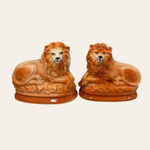 May include: Two ceramic lion figurines in a reclining position, each featuring a glossy orange-brown finish, detailed manes, and painted facial features. The lions are resting on oval-shaped bases, showcasing a smooth, reflective surface texture.