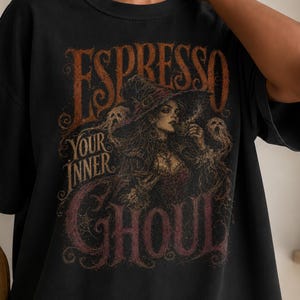 Può includere: Una t-shirt nera a maniche corte con una stampa grafica raffigurante una strega con capelli lunghi e un cappello, circondata da figure spettrali. Il testo "ESPRESSO YOUR INNER GHOUL" è scritto in un carattere stilizzato e usurato nei toni dell'arancione.