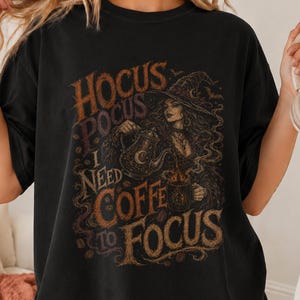 May include: A black short-sleeve t-shirt featuring a graphic print of a witch pouring coffee from a teapot into a mug. The text reads HOCUS POCUS I NEED COFFEE TO FOCUS in a stylised brown and orange font with swirling steam and small bats.