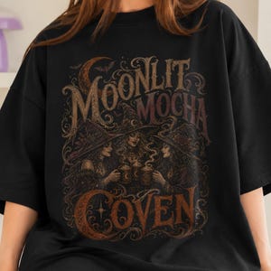 May include: A black t-shirt featuring a graphic print with the text "MOONLIT MOCHA COVEN". The design depicts three figures wearing pointed hats, holding steaming mugs, surrounded by intricate swirling patterns and a crescent moon in brown tones.