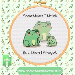 Pode incluir: Um padrão de ponto cruz num bastidor de madeira com três sapos verdes de bochechas rosadas. O texto diz: Sometimes I think, But then I froget. O design é exibido num fundo de tecido branco com um padrão de folhas verde-claras.