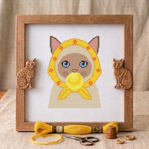 May include: A cross-stitch embroidery of a cat wearing a yellow floral headscarf and blowing a yellow bubble, framed in a wooden square frame with two carved wooden cat figures attached to the sides, accompanied by yellow embroidery thread.