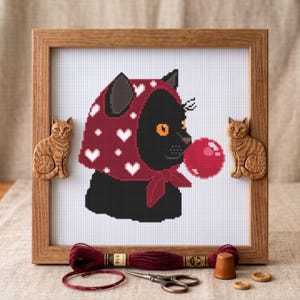 Pode incluir: Um bordado em ponto cruz de um gato preto usando um lenço vermelho escuro com corações brancos, soprando uma bolha rosa. A peça está numa moldura de madeira com duas pequenas figuras de gato de madeira nas laterais, acompanhada de fio.
