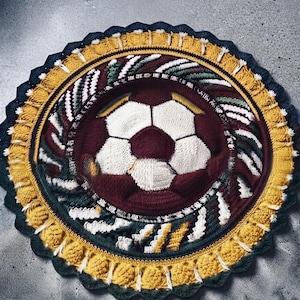 May include: A circular crochet rug featuring a central soccer ball motif in white and dark red. The design is surrounded by concentric rings of textured patterns in dark green, white, and yellow, finished with a scalloped dark blue border.