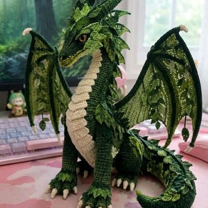 Op de afbeelding: Een gehaakt drakenfiguur in tinten donker- en lichtgroen garen. Het wezen heeft een crèmekleurige buik, bladachtige schubben langs de rug en vleugels, en witte klauwen. Het staat in een viervoetige houding met de vleugels wijd uitgespreid.