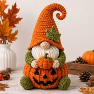 Peut inclure: Une figurine de gnome au crochet avec un grand chapeau orange recourbé orné d'une feuille verte. Le gnome possède une barbe en laine blanche, des manches vert olive et repose sur une base en forme de citrouille orange avec un visage brodé.