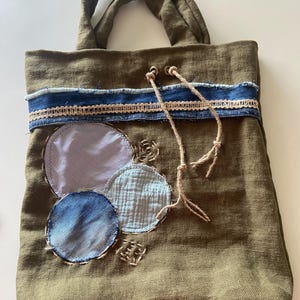 Peut inclure: Un sac fourre-tout vert olive orné d'une bande horizontale de tissu denim bleu avec une bordure en dentelle beige. L'avant est décoré de trois pièces de tissu circulaires bleues et blanches, ainsi que de deux cordons en jute pendants.