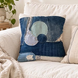 Peut inclure: Un coussin carré composé d'un assemblage de tissus en jean bleu foncé. La surface présente des appliqués circulaires en tissu bleu clair et bleu sarcelle, agrémentés de broderies faites à la main, de bords effilochés et de petits motifs noirs.