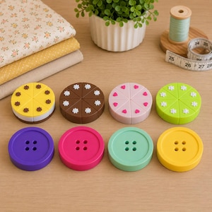 Puede incluir: Una colección de ocho objetos circulares dispuestos en dos filas. La fila superior presenta cuatro objetos con forma de pastel en amarillo, marrón, rosa y verde, divididos en segmentos con pequeños detalles florales o de corazones. La fila inferior contiene cuatro botones de colores sólidos en morado, rosa intenso, verde menta y amarillo, cada uno con cuatro agujeros centrales.