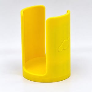 Puede incluir: Un portavasos cilíndrico de color amarillo brillante con una amplia abertura vertical en un lado. El soporte cuenta con una base circular sólida y un acabado mate liso con un pequeño logotipo abstracto en relieve en el exterior.