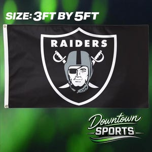 May include: A black rectangular flag featuring the Raiders logo, which consists of a shield with a pirate wearing an eye patch and two crossed swords. The text SIZE: 3FT BY 5FT is printed above the flag, and the text Downtown SPORTS is below it.