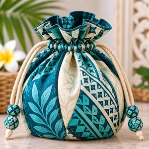 Könnte beinhalten: Ein runder Stoffbeutel mit Kordelzug, der abwechselnde Paneele in Blaugrün und Creme mit botanischen Blattmustern und geometrischen Formen aufweist. Der Beutel wird mit dicken cremefarbenen Kordeln gerafft, die mit je zwei passenden Perlen enden.