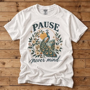 Könnte beinhalten: Ein weißes kurzärmeliges T-Shirt mit einem Grafikdruck eines Pfaus, umgeben von floralen Elementen und einer Sonne. Der Text PAUSE ist in Dunkelblau über dem Pfau gedruckt, und der Text never mind... ist in Dunkelblau darunter gedruckt.
