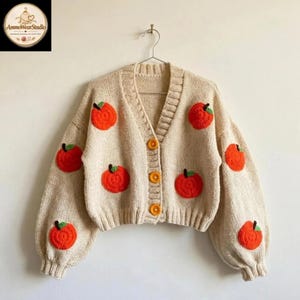 Op de afbeelding: Een beige gebreid vest met een V-hals, lange mouwen met geribbelde boorden en drie oranje knopen. Het kledingstuk is versierd met verschillende gehaakte oranje fruitapplicaties, elk voorzien van een klein groen blaadje.