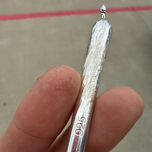 May include: A silver-colored cylindrical object with a tapered end, featuring an engraved leaf pattern along the body. The surface is stamped with the number 925 and a small circular hallmark near the base, indicating sterling silver composition.
