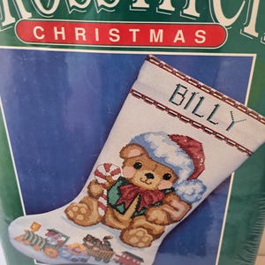 May include: A Bernat cross stitch kit for a Christmas stocking featuring a teddy bear wearing a Santa hat and holding a candy cane. The stocking is personalized with the name BILLY and includes a toy train design at the bottom.