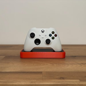 Puede incluir: Un mando de videojuegos inalámbrico blanco con dos joysticks negros, una cruceta y cuatro botones de acción de colores etiquetados como A, B, X e Y, apoyado sobre una base de carga rectangular de color negro y naranja.