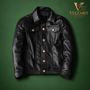 May include: A black leather jacket featuring a pointed collar, long sleeves, and a front button closure with metallic gold-toned buttons. Two chest pockets with buttoned flaps are visible. The logo VELZARO LEATHERS is printed in the top right corner.