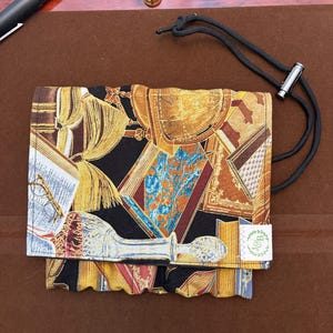 May include: A fabric roll-up case featuring a black background with a print of vintage books, a globe, and a glass decanter. The case includes a black cord with a metal toggle for closure and a small white tag with the text J&B in a green wreath.