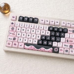 Puede incluir: Un teclado mecánico con teclas de color rosa claro y negro. Varias teclas muestran pequeñas ilustraciones de personajes de dibujos animados. La barra espaciadora tiene un patrón ondulado rosa y negro con puntos. Incluye teclas QWERTY.