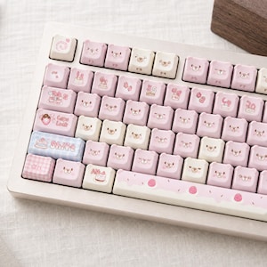 May include: A mechanical keyboard featuring custom keycaps in shades of cream and pastel pink. The keys are shaped like bear heads, some with printed bear faces, cakes, and sweets. The shift key is blue plaid, and the spacebar has pink frosting.
