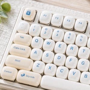 May include: A computer keyboard featuring rounded, glossy off-white keycaps with blue lettering. The keys include standard QWERTY characters, function keys, and custom icons such as a jar with a heart and the text Soy milk on the bottom left key.