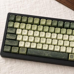 May include: A mechanical keyboard featuring a color scheme of sage green and cream keys. Several keys, including the escape, tab, caps lock, shift, and spacebar, display intricate botanical and floral patterns in a darker forest green tone.