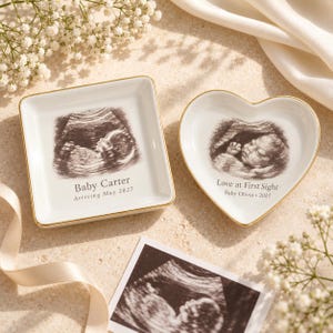 May include: A square white ceramic trinket dish with gold trim featuring a black and white ultrasound image, the text Baby Carter, and Arriving May 2027, alongside a heart-shaped dish with an ultrasound, Love at First Sight, and Baby Olivia 2027.