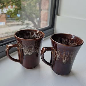May include: Two ceramic mugs featuring a glossy dark brown glaze with a distinctive white drip pattern around the upper rim. Each mug has a flared opening, a rounded body, and a sturdy, curved handle extending from the side of the vessel.