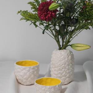 Könnte beinhalten: Ein Set aus drei Keramikgefäßen, bestehend aus einer hohen Vase und zwei kleineren Schalen. Jedes Stück hat eine weiße, texturierte, unebene Außenfläche und ein glattes, leuchtend gelbes Inneres. Die Vase enthält einen Blumenstrauß.