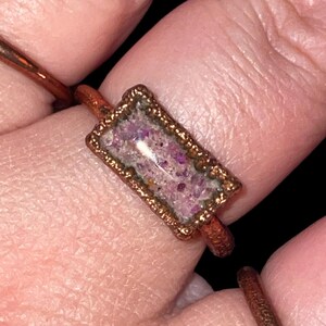 May include: A handcrafted copper ring with a rectangular, bezel-set stone. The stone is filled with small, sparkling purple and pink fragments. The ring has a rustic, artisan look, with a dark background.