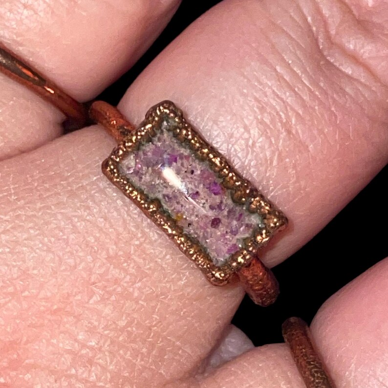 May include: A handcrafted copper ring with a rectangular, bezel-set stone. The stone is filled with small, sparkling purple and pink fragments. The ring has a rustic, artisan look, with a dark background.