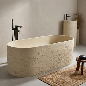 Puede incluir: Una bañera exenta de forma ovalada con un acabado de piedra beige texturizada. La bañera cuenta con un grifo de suelo negro mate con ducha de mano. Un lavabo de pedestal cilíndrico de piedra a juego se encuentra cerca con un grifo negro.