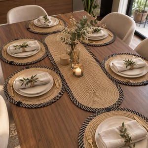 May include: A dining table setting featuring a long oval woven jute table runner and six matching round placemats with black and natural tan braided edges. Each place setting includes a white ceramic plate, a folded linen napkin, and gold cutlery.