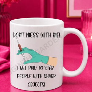 May include: A white ceramic mug featuring a graphic of a hand in a teal medical glove holding a syringe filled with red liquid. The text reads: DON'T MESS WITH ME! I GET PAID TO STAB PEOPLE WITH SHARP OBJECTS! The mug has a curved handle on the right.
