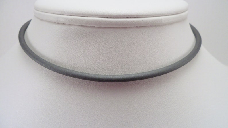 May include: A simple, dark gray choker necklace made of a smooth, flexible material. The necklace is shown on a white mannequin.
