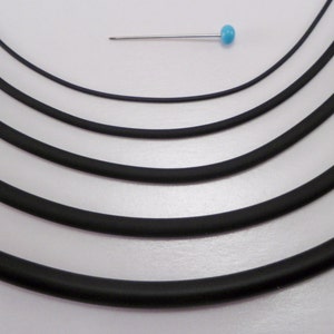 May include: Five different sizes of black elastic cord, arranged in a circle on a white background. A small blue pin is placed above the cords for size reference.