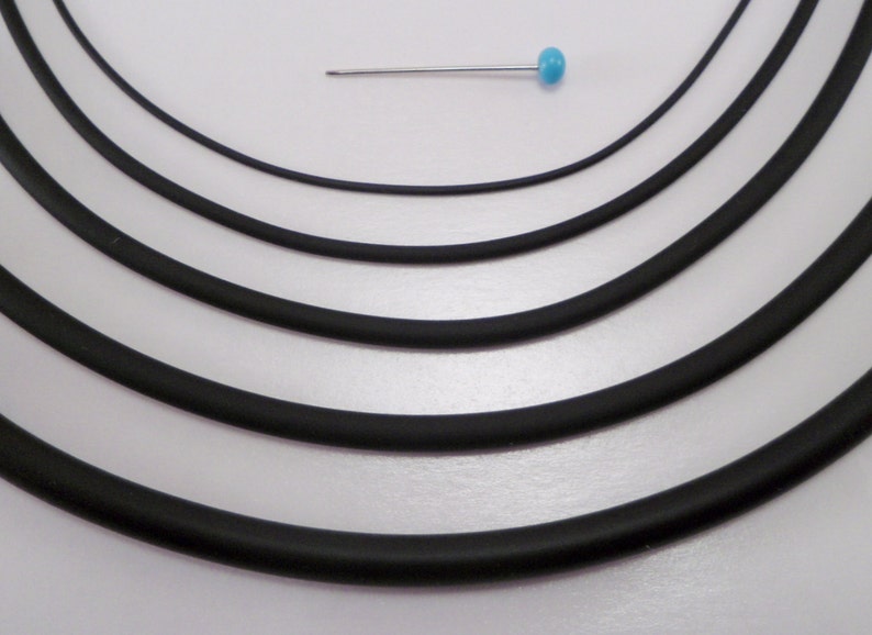 May include: Five different sizes of black elastic cord, arranged in a circle on a white background. A small blue pin is placed above the cords for size reference.