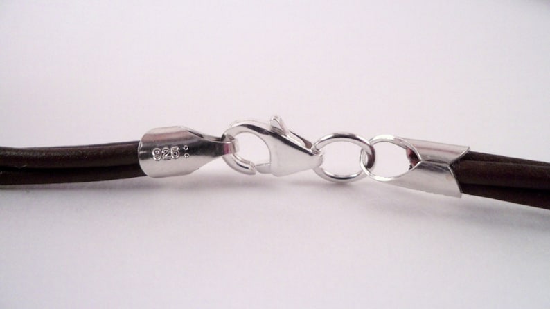 May include: Close-up of a brown leather cord necklace with a silver clasp. The clasp is marked with "925."