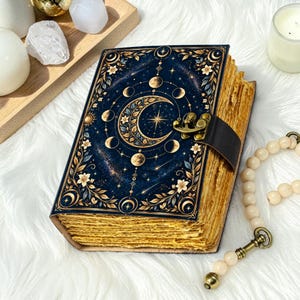 May include: A rectangular journal with a dark blue cover featuring gold celestial moon phases, stars, and floral patterns. The book has a dark brown leather strap closure with a brass latch and thick, deckled-edge pages in a golden tan color.