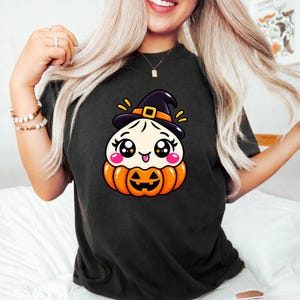 Könnte beinhalten: Ein schwarzes T-Shirt mit einer Grafik eines weißen Knödel-Charakters, der einen schwarzen Hexenhut mit orangefarbenem Band trägt und in einem orangefarbenen Kürbis sitzt. Die Figur hat große funkelnde Augen, rosa Wangen und eine kleine Zunge.