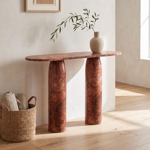 Può includere: Un tavolo console con un piano ovale allungato e due gambe cilindriche spesse, caratterizzato da una ricca venatura del legno radica color bruno-rossastro. Sul piano del tavolo poggia un vaso in ceramica beige con un ramo d'ulivo verde.