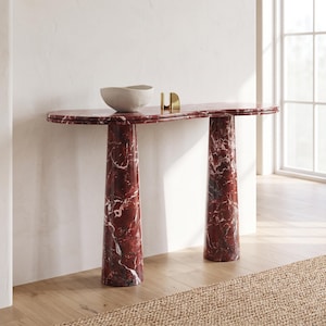 May include: A console table crafted from deep red marble with white veining, featuring an elongated oval top supported by two cylindrical pedestal legs. A matte white bowl and a small gold-toned metal object sit on the table surface.